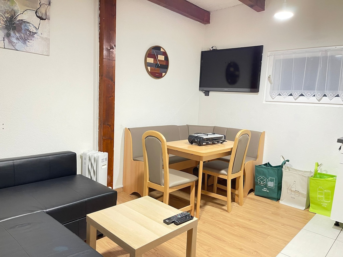 A modern living area is depicted, featuring a black sofa and a light wood dining table surrounded by chairs. A flat-screen television is mounted on the wall, while art and a circular decorative piece adorn the space. Natural light is provided by a nearby window.