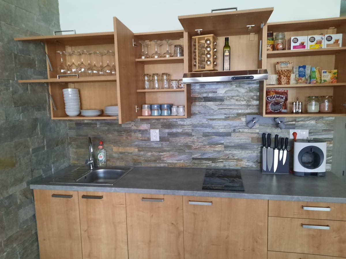The kitchen features a blend of wooden cabinets and a stone backsplash. Shelves hold an assortment of glasses, plates, and jars, while a sink and cooktop are integrated into the countertop. A knife block and dishwashing machine are also visible, promoting convenience during meal preparation.