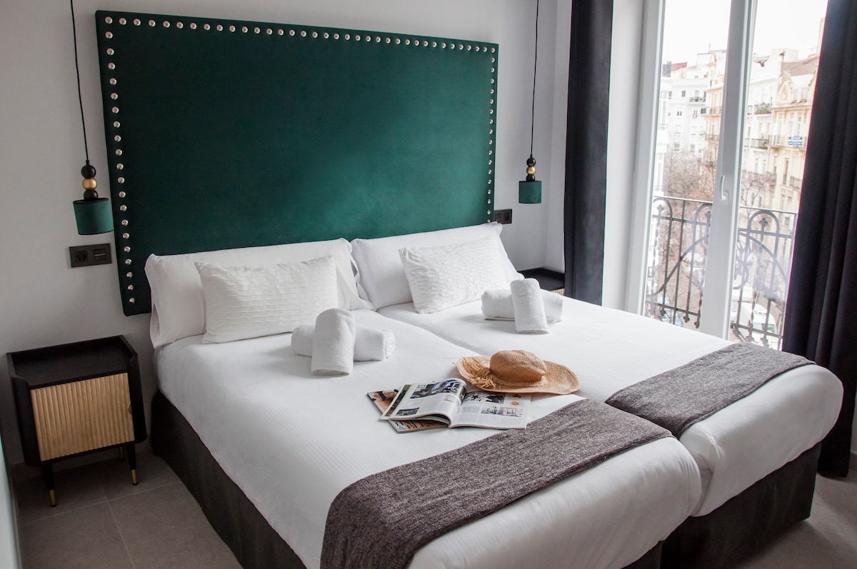 A room featuring two spacious beds with crisp white linens and decorative headboards. Soft towels are neatly arranged on the beds. A magazine and a straw hat are placed on a bedside table, with large windows offering natural light and views of the surrounding area.