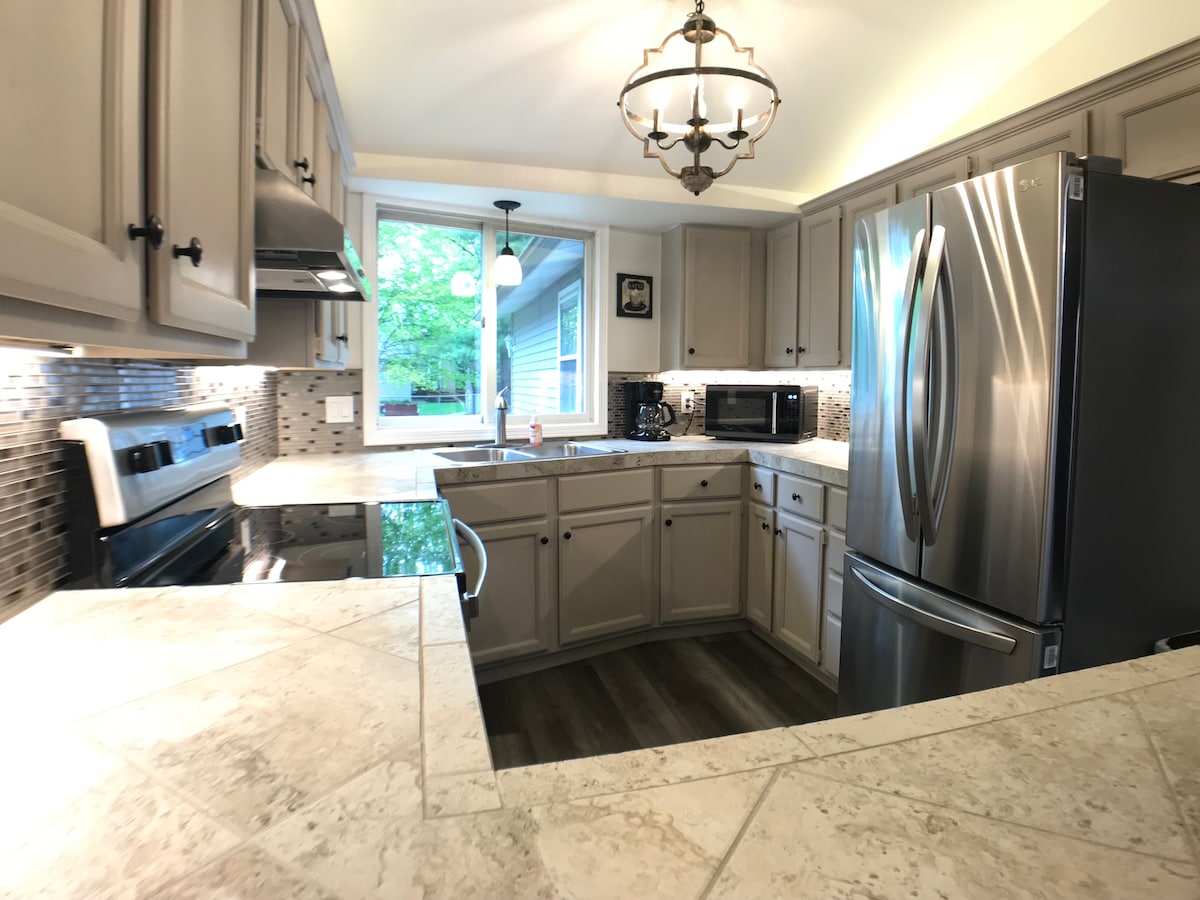 The kitchen features a modern design with light gray cabinetry and stainless steel appliances. A spacious countertop made of marbled stone allows for ample workspace. Soft lighting is provided by a chandelier overhead, while natural light enters through a window above the sink.