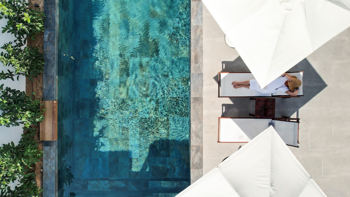 An aerial view of a luxurious rectangular pool surrounded by shaded lounge areas. Two sunbeds are placed nearby, with sun umbrellas providing cover. The clear water reflects the surrounding greenery, creating a serene environment within the outdoor space.