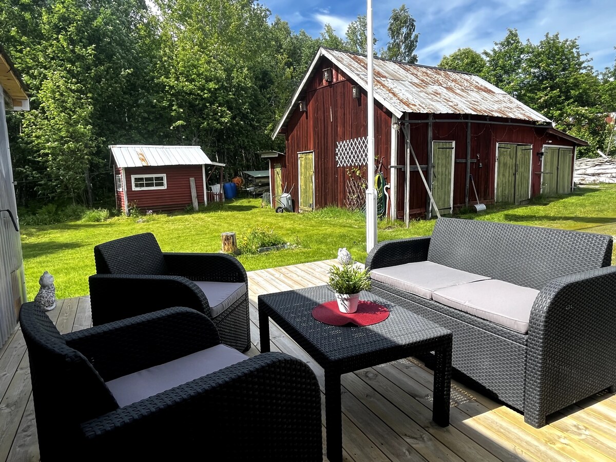 An outdoor lounge area is furnished with black wicker seating and a small table, set on a wooden deck. Lush greenery surrounds the space, with a red barn visible in the background, complemented by a clear blue sky.