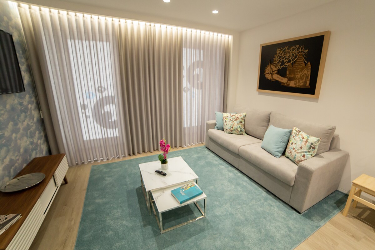 A well-lit living area features a soft grey sofa adorned with decorative pillows. A coffee table sits in front, accompanied by a light blue area rug. Two large windows dressed in sheer curtains allow natural light to filter in, enhancing the inviting space.
