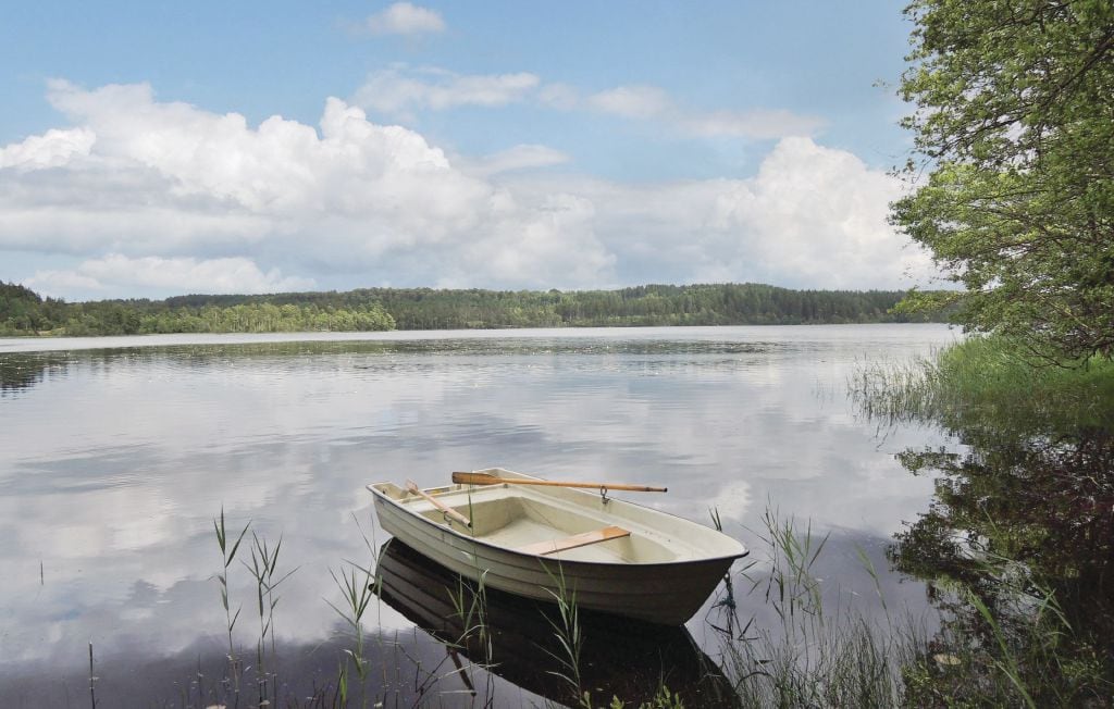 A small white boat rests gently on calm waters, surrounded by lush trees and distant greenery. The sky is dotted with fluffy clouds, reflecting softly on the lake's surface, while tall reeds frame the scene, adding to the natural tranquility.