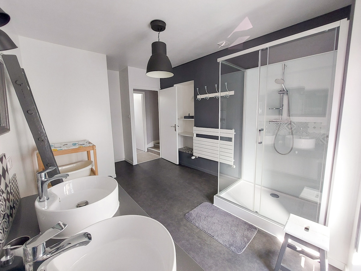 A spacious bathroom is equipped with two sinks and a large shower enclosure. Natural light fills the area, illuminating the neutral-toned walls and grey flooring. A towel rack and a small stool are present, contributing to the functionality of the room.