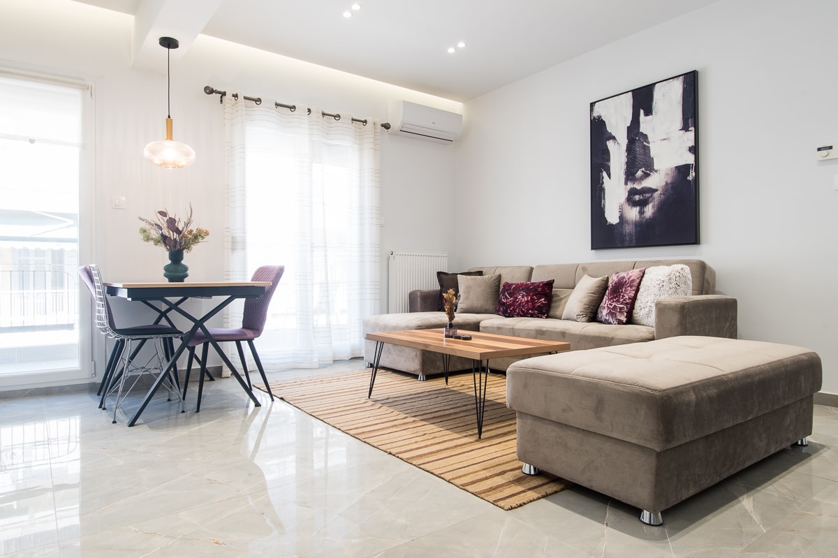 A spacious living area is defined by a modern sofa in neutral tones, accented by colorful cushions. A wooden coffee table and a dining set are positioned comfortably, with natural light filtering through sheer curtains. A wall-mounted artwork complements the contemporary decor.