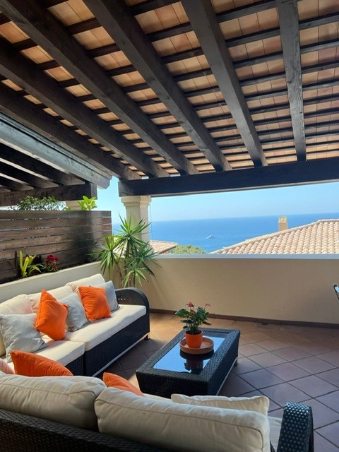 A covered outdoor seating area features a comfortable sofa adorned with orange cushions. A small table sits in front, accompanied by a potted plant. The space is enhanced by views of the sea and surrounding landscape, with wooden beams visible in the ceiling.