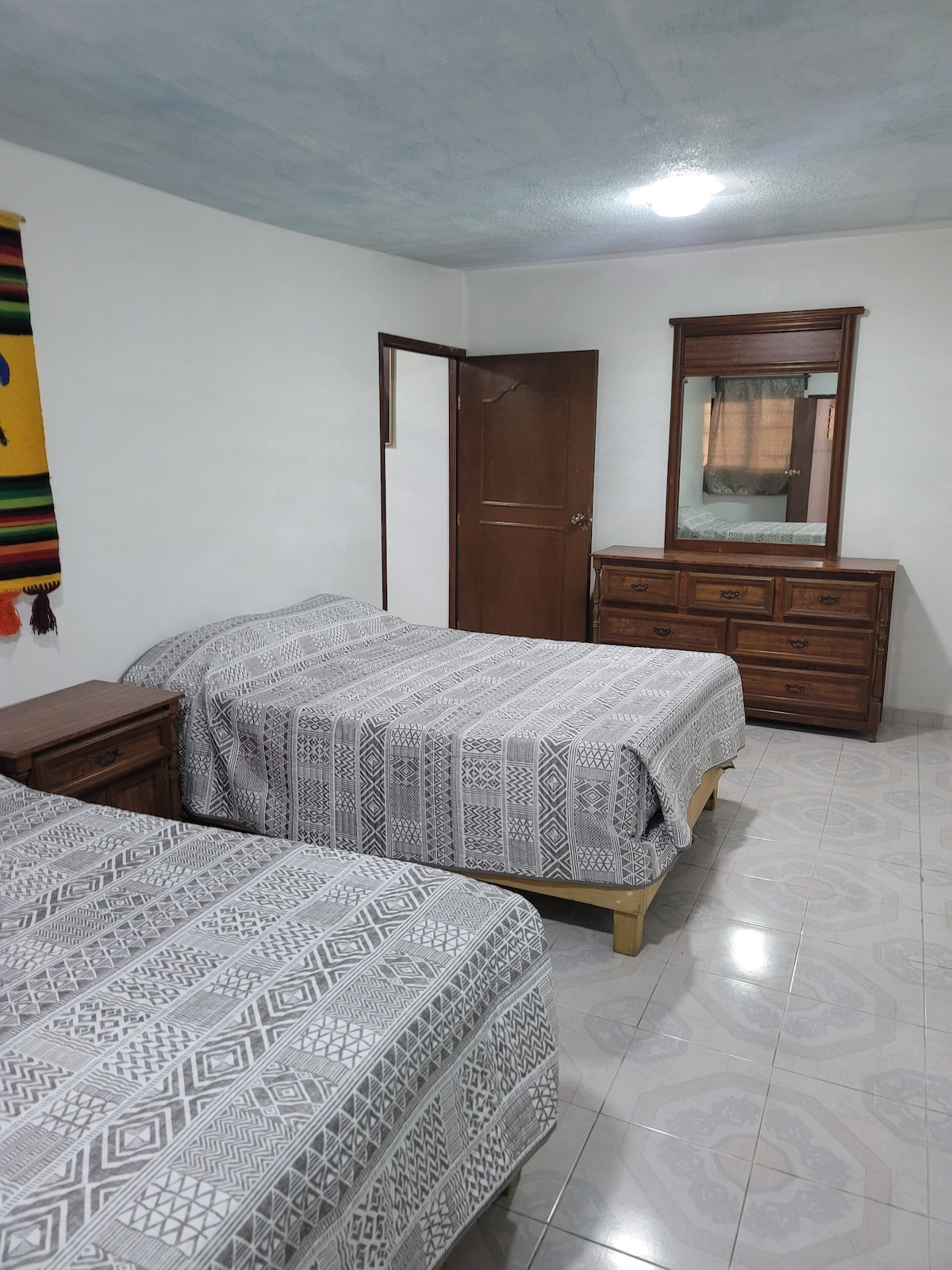 A well-appointed sleeping area features two spacious beds with patterned blankets. A wooden dresser and mirror are positioned against the wall, while a door leads to another room. Soft lighting and tiled flooring create a comfortable environment.