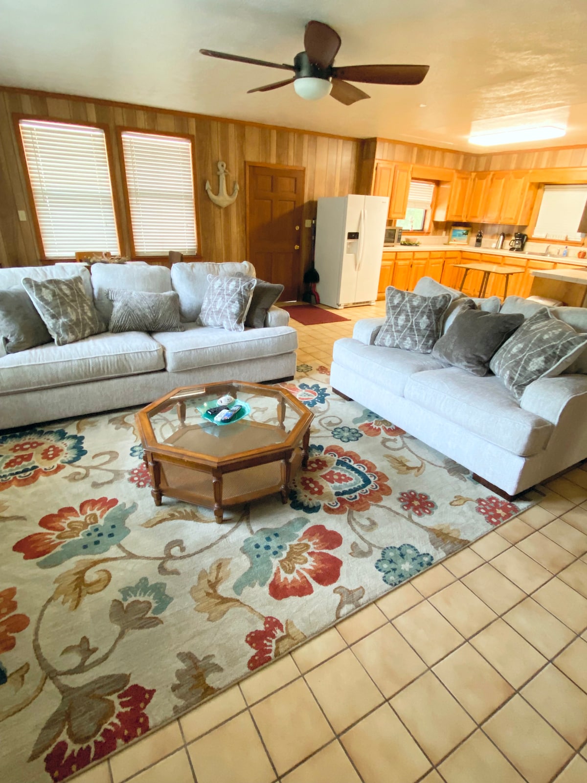 A welcoming living area features two comfortable sofas adorned with decorative pillows. A wooden coffee table is positioned at the center, resting on a large, colorful area rug. The space is brightened by natural light from multiple windows, and warm wood paneling complements the setting.