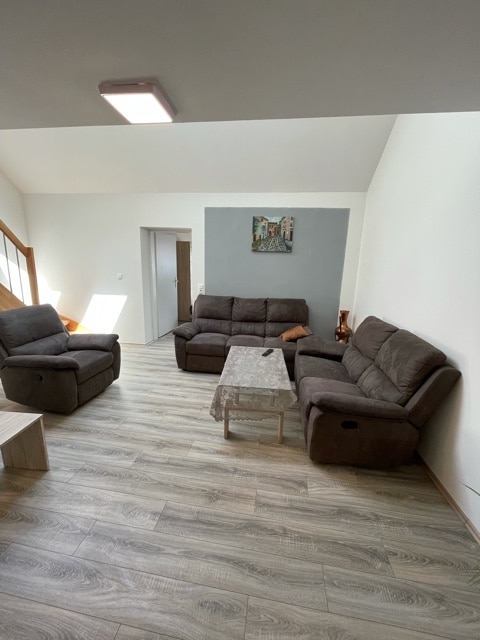 A spacious living area features two comfortable dark gray sofas positioned around a light wooden coffee table. A piece of art hangs on the wall, and natural light filters in from a nearby window, enhancing the inviting environment.
