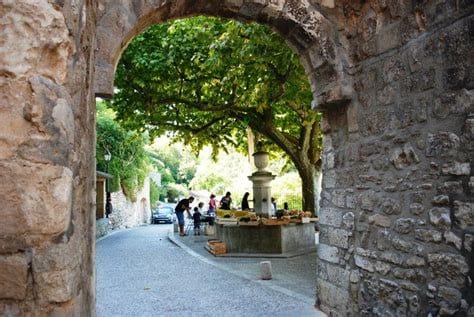 A view through a stone archway reveals a tree-lined street bustling with activity. A group of people gathers around a market stall filled with goods, while a few cars are parked along the road. The greenery above provides shade, enhancing the lively atmosphere.