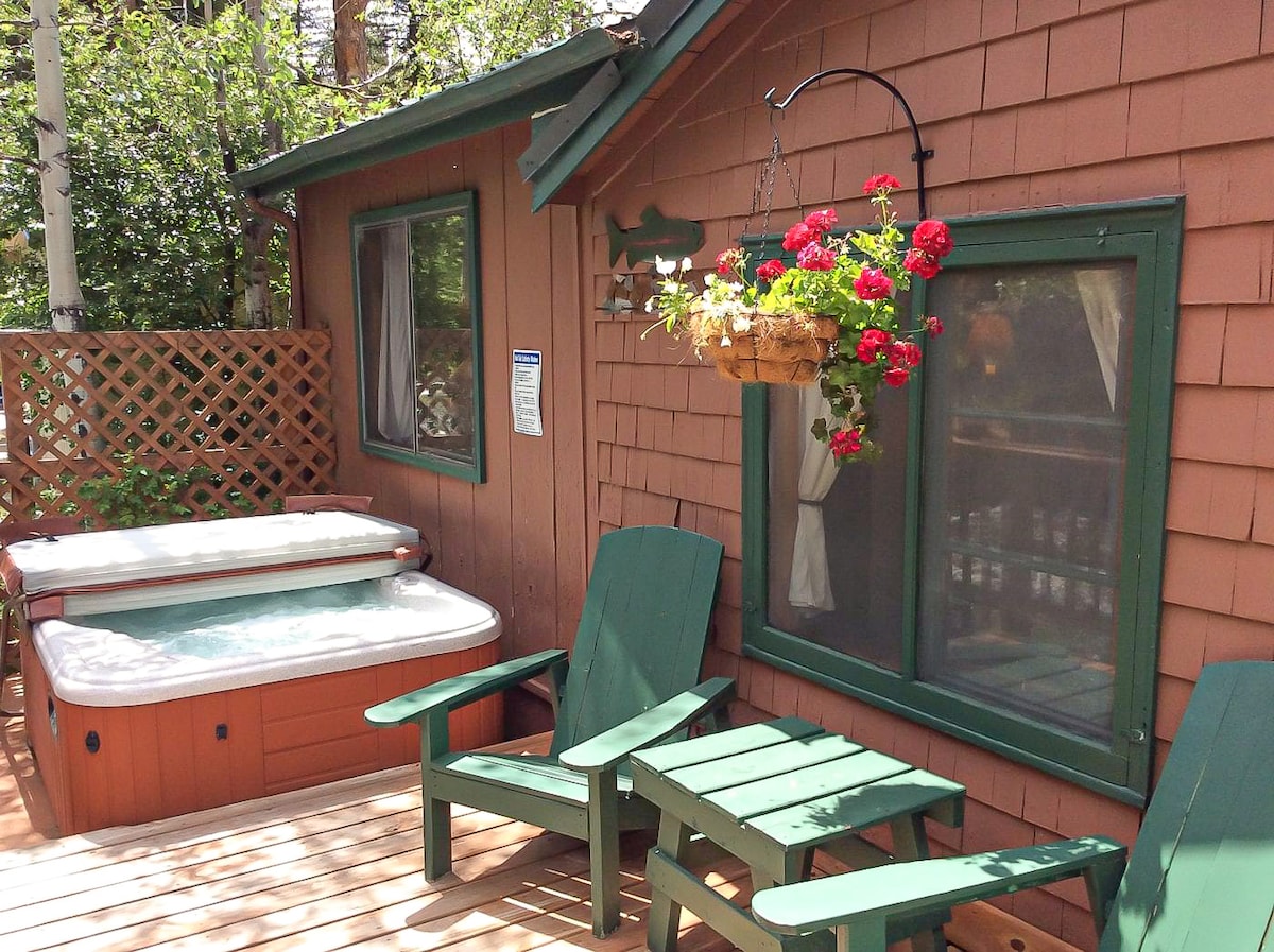 A wooden deck features two green Adirondack chairs positioned beside a hot tub. A hanging flower pot filled with vibrant blossoms adds color against the rustic brown siding of the cabin. Windows are framed in green, complementing the natural surroundings.