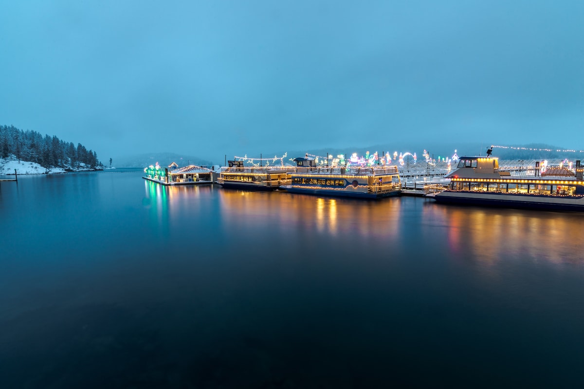 A panoramic view of a tranquil lake at dusk, reflecting soft lights from various structures along the waterfront. Snow-dusted trees line the shore, while colorful decorations create a festive ambiance across the water, enhancing the serene atmosphere of the scene.