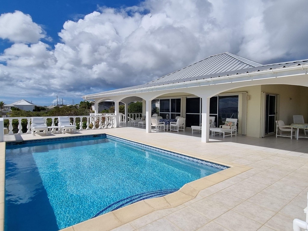 A clear blue pool reflects the sky, framed by a white tiled patio and adjacent lounge chairs. Spacious overhangs provide shade for outdoor seating areas. The villa's open architecture allows for easy access to both the pool and the surrounding views of the landscape.