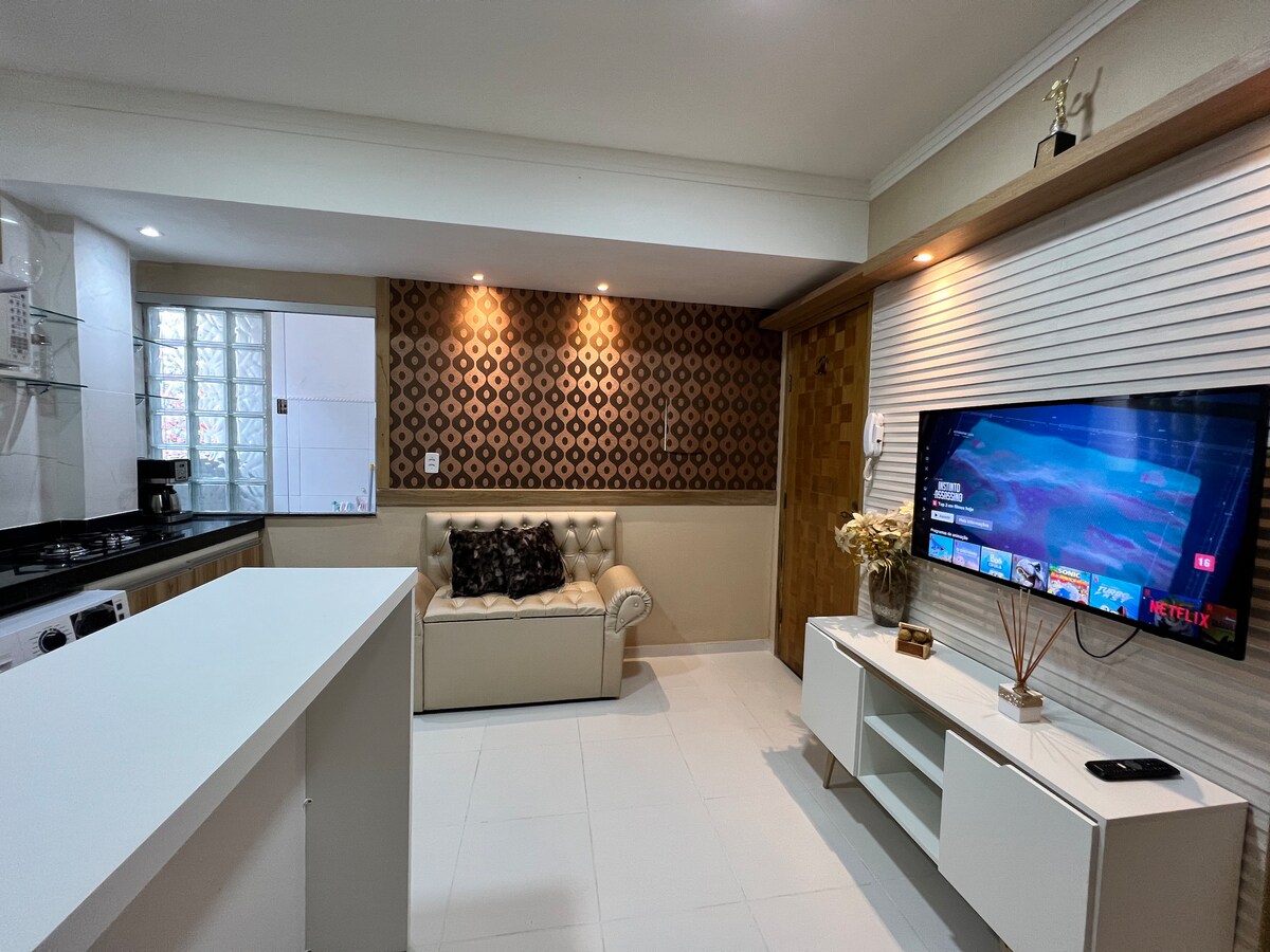 A modern living area is highlighted by a white couch with black accents. A sleek television is mounted on the wall, accompanied by a light wooden media console. The kitchen area features clean lines and ample counter space, with a glass block window allowing natural light.