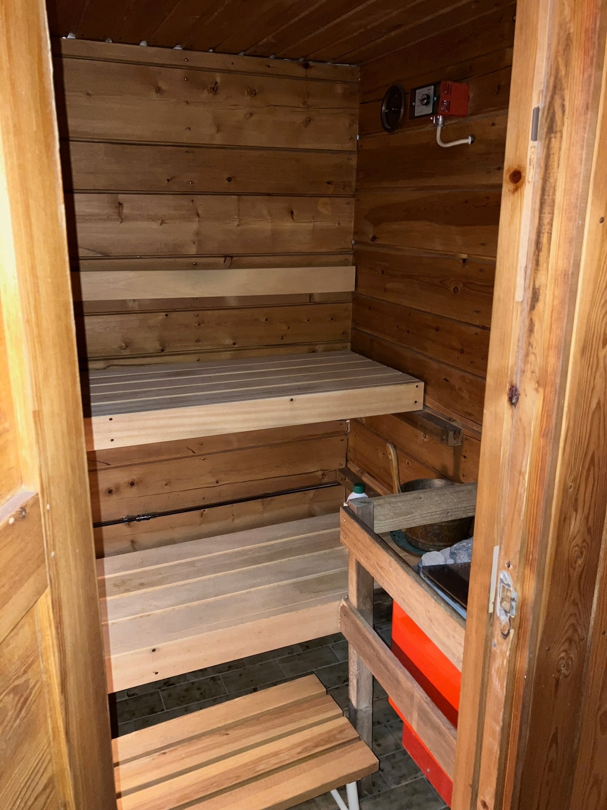 A traditionally designed sauna interior features wooden walls and benches. Several shelves are available for relaxation, and a small stool is positioned at the side. Natural light reflects softly on the polished wood, enhancing the warm feel of the space.