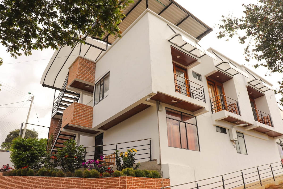 A modern multi-level building is showcased, featuring a blend of white and brick exteriors. Large windows are framed by wooden accents, allowing natural light to fill the space. Colorful flowers line the entrance, adding a touch of nature to the contemporary design.