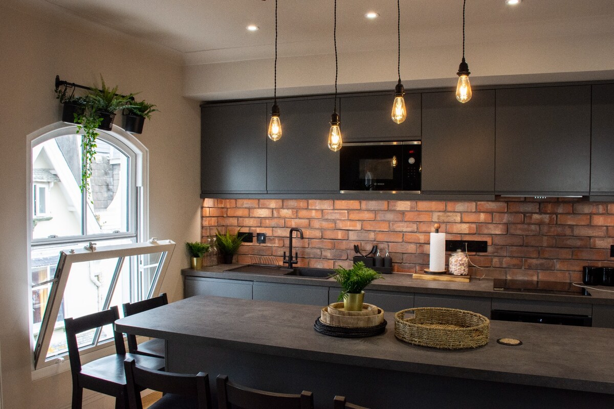 The kitchen is equipped with modern appliances and features a dark cabinetry contrasted by a rustic brick backsplash. Overhead, pendant lights illuminate the counter that includes a bowl of greenery and a decorative tray. A window allows for natural light and fresh air.
