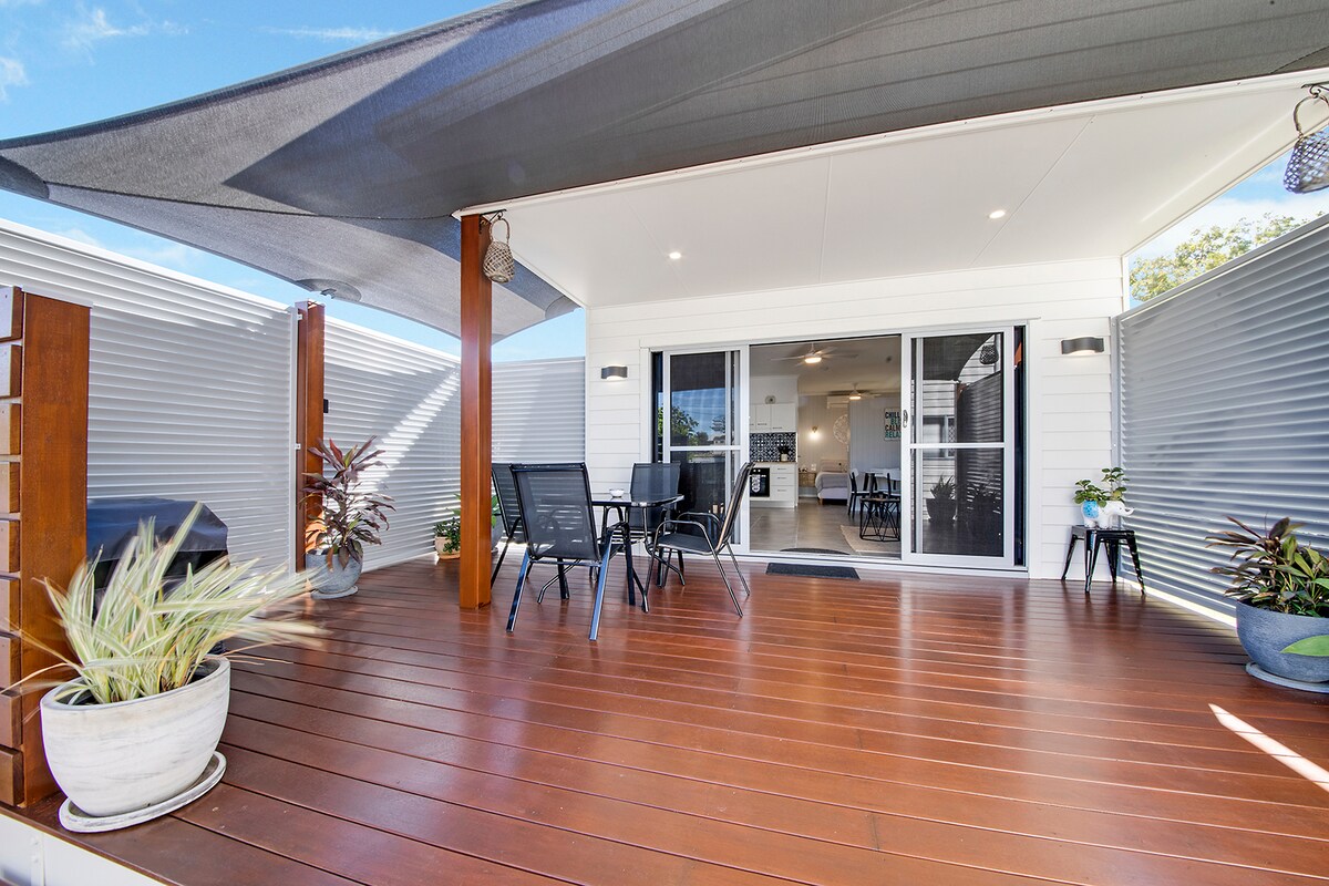 An outdoor deck features wooden flooring with several black chairs surrounded by potted plants. The area is shaded by a large canopy, and sliding glass doors lead into the interior living space. Bright light filters through, creating an inviting ambiance.
