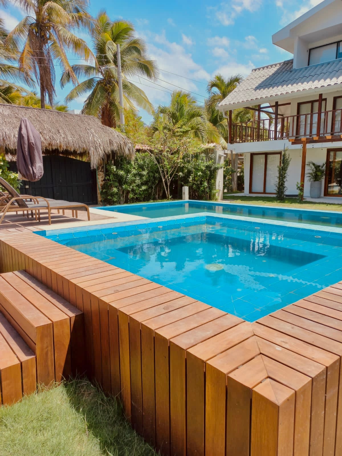 A refreshing swimming pool is highlighted, surrounded by wooden decking and lush tropical greenery. A thatched-roof structure provides shade nearby, while loungers are positioned for relaxation. The clear water reflects the blue sky and creates a serene outdoor space for guests.