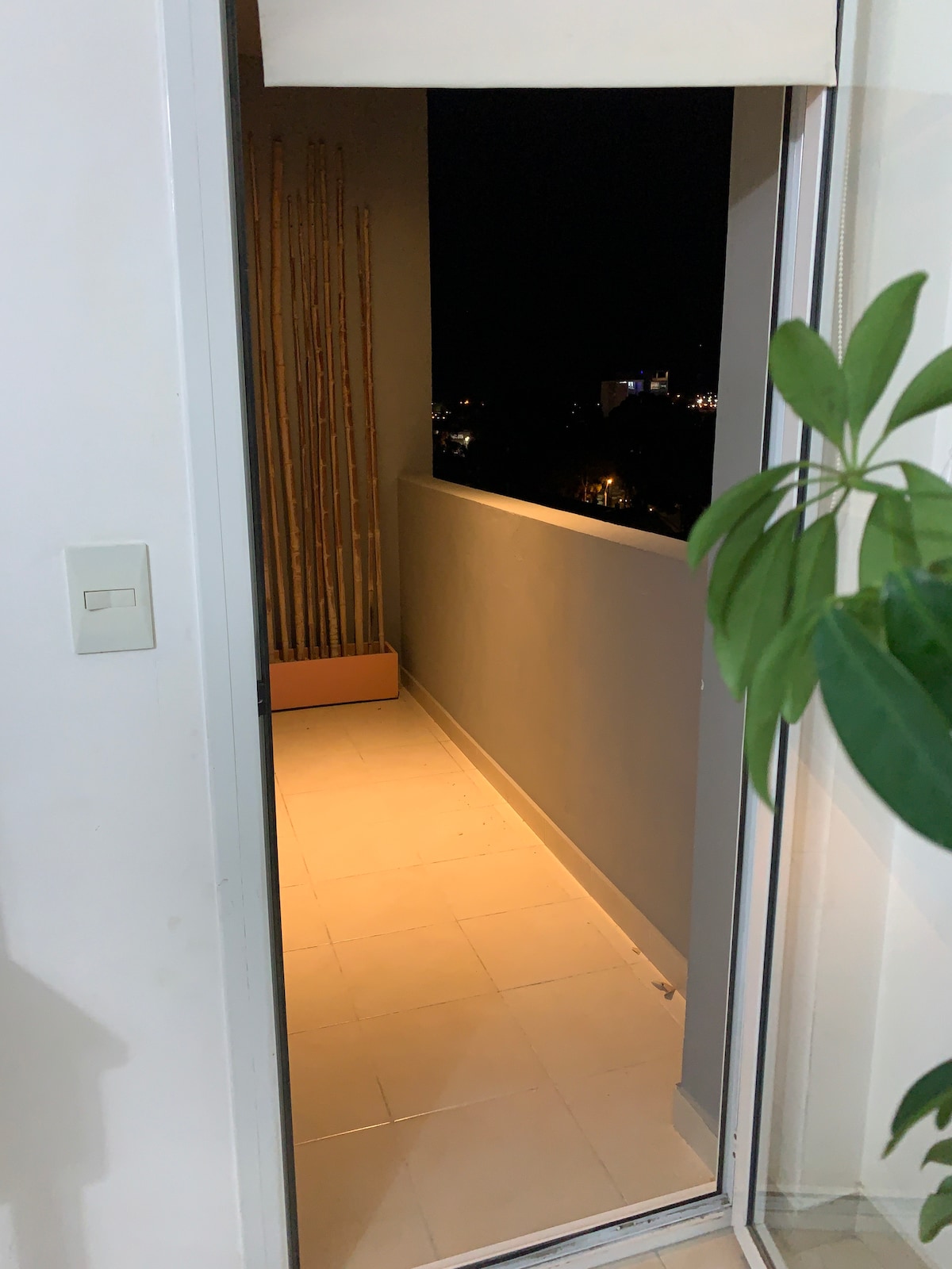 A private balcony area is illuminated softly, featuring a sleek tile floor and a minimalist plant near the entrance. The view includes distant city lights visible through the open doorway, providing a serene transition from indoors to outdoors.