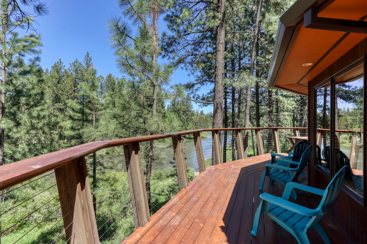 A spacious deck surrounds the round cabin, featuring several teal chairs positioned for relaxation. Tall pine trees frame the view, with the Spring River visible in the distance, providing a serene, natural backdrop.