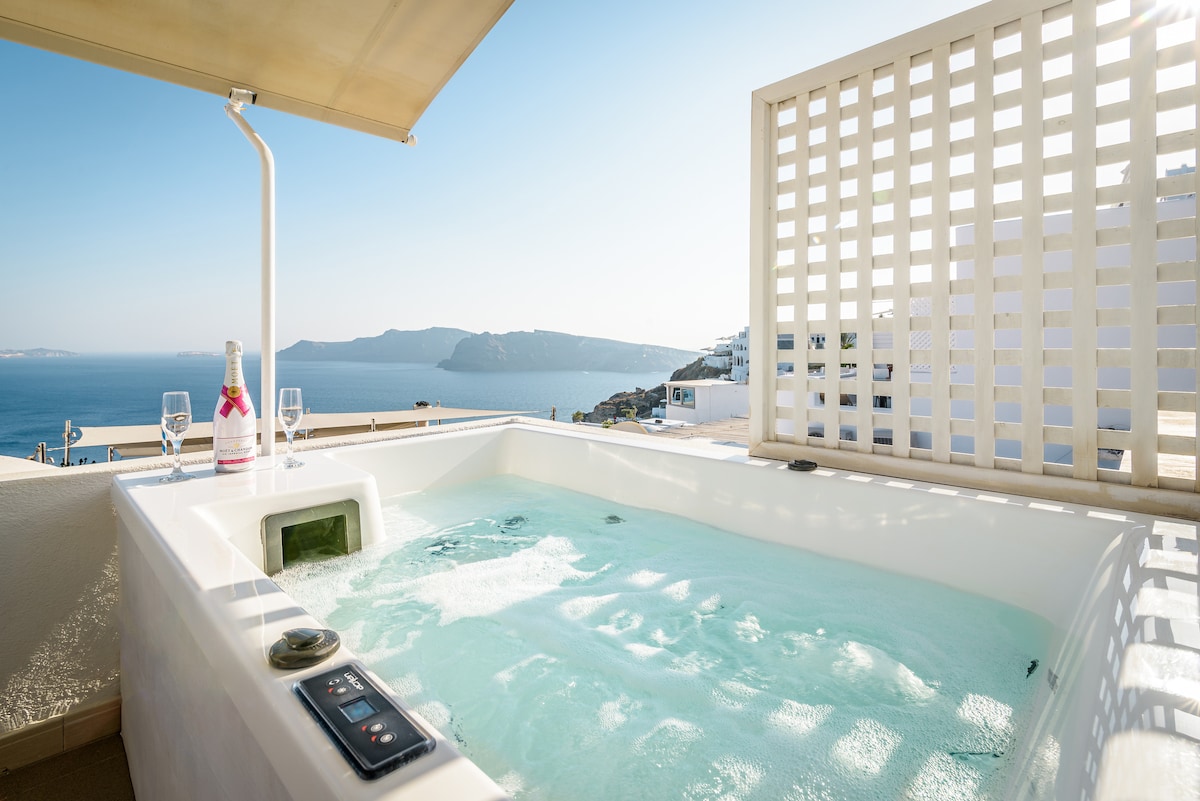 A private jacuzzi is positioned on a balcony overlooking the serene caldera and distant cliffs. Clear water gently ripples, while two glasses and a bottle of champagne rest on the nearby ledge, enhancing the tranquil setting with stunning panoramic views.