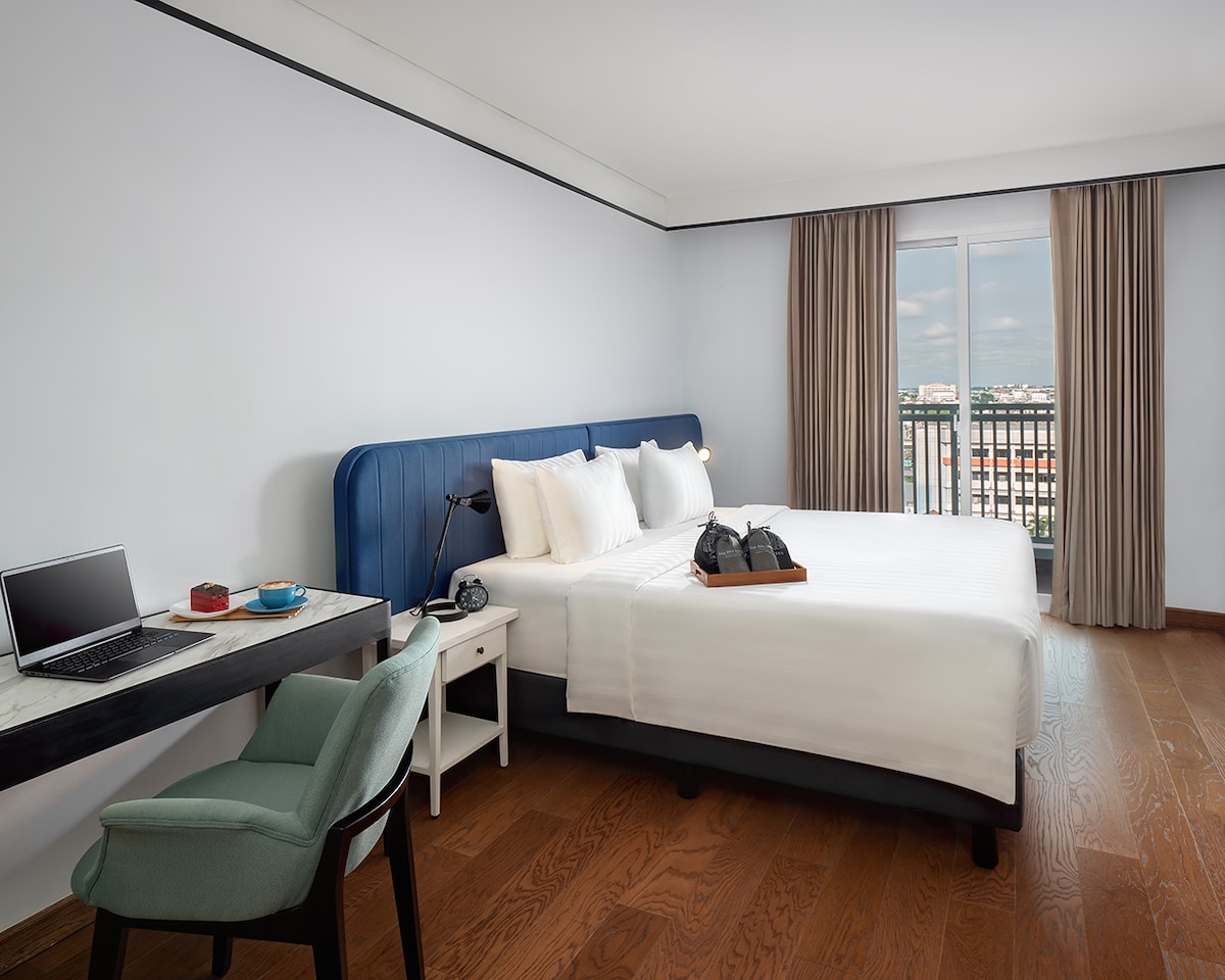 A modern bedroom features a spacious bed with white linens against a blue headboard. A small desk with a laptop and a chair is positioned nearby, while large windows with light curtains allow natural light to enter the room.