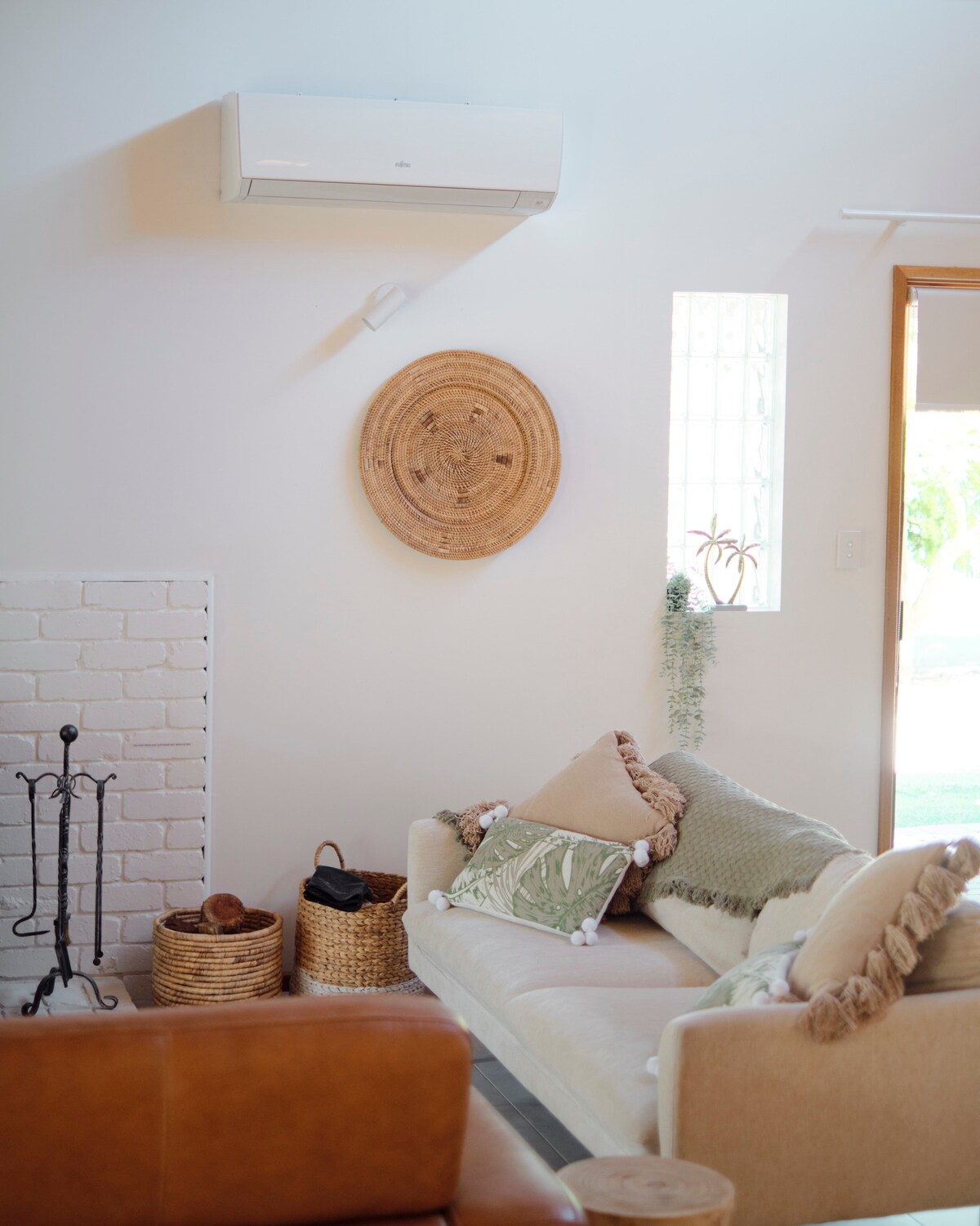 A light-filled living space features a comfortable sofa adorned with various textured pillows. Natural decor elements are present, including a woven wall hanging and a nearby door opening to the outdoors. Air-conditioning is installed for climate control, enhancing the room's comfort.