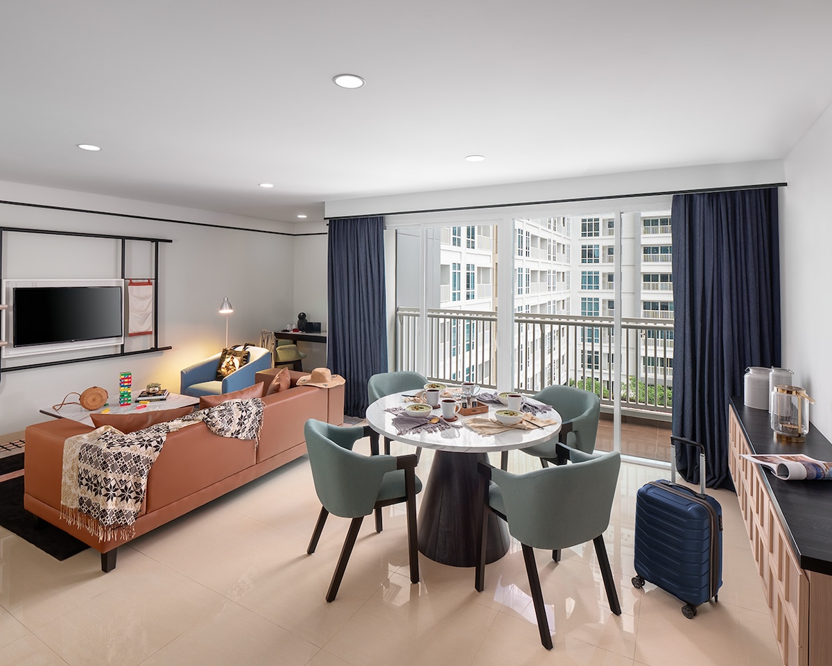 A modern living space features a comfortable couch adorned with a throw and cushions. A round dining table with four chairs is set nearby. Large windows allow natural light, with sheer curtains partially drawn. A media unit holds a television, adding to the functionality of the area.