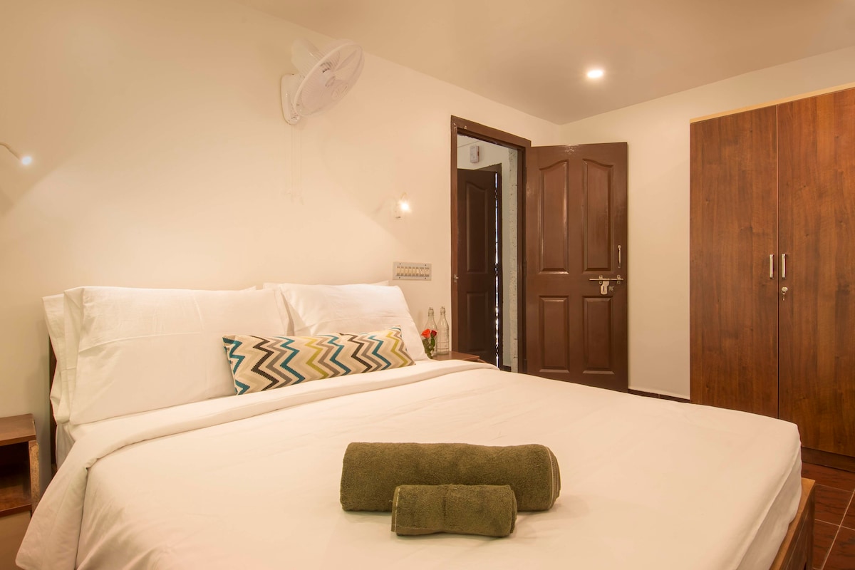 A spacious bedroom is presented with a comfortable bed featuring a light-colored duvet and a decorative pillow with geometric patterns. A set of clean towels is neatly placed at the foot of the bed. The room contains a wardrobe and features warm, inviting lighting.