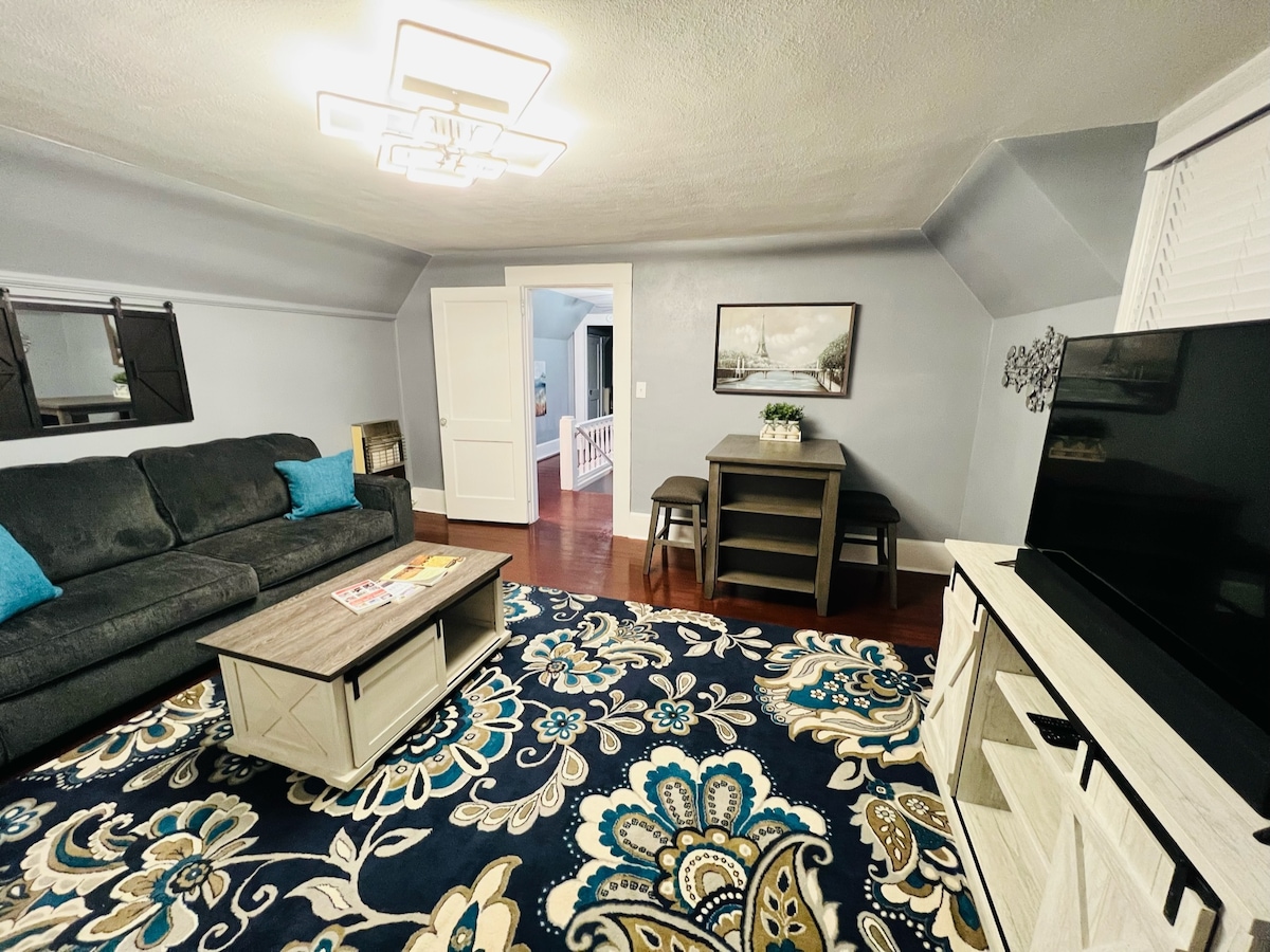 A spacious living area showcases a plush dark sofa with blue accent pillows, paired with a coffee table adorned with magazines. A large area rug featuring intricate patterns covers the hardwood floor, and a flat-screen TV is positioned on a light-colored media console. Natural light enters through multiple windows.