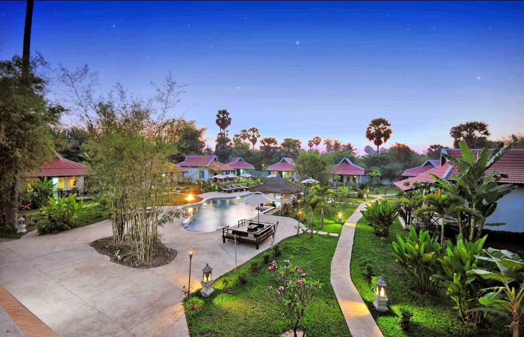 A picturesque outdoor landscape is revealed, featuring a serene pool surrounded by lush greenery and colorful flowers. Pathways meander through the gardens, leading to modern-style accommodations with sloped roofs. Ambient lighting enhances the overall tranquil ambiance as dusk settles in.