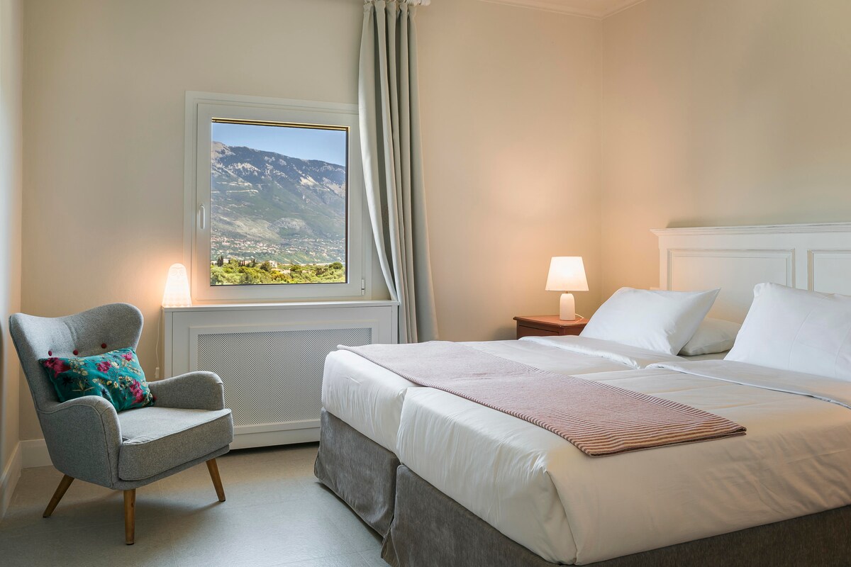 A well-lit bedroom features a spacious bed with light-colored bedding and a soft throw. A grey armchair with a decorative pillow occupies one corner. A window offers scenic views of the mountains, while a bedside table with a lamp provides additional lighting.