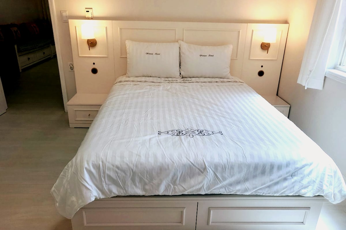 A spacious bed with a white quilted coverlet is positioned against a light-colored wall. Soft bedside lamps are affixed on either side of the headboard, creating a serene atmosphere. Natural light filters through a nearby window with light curtains.