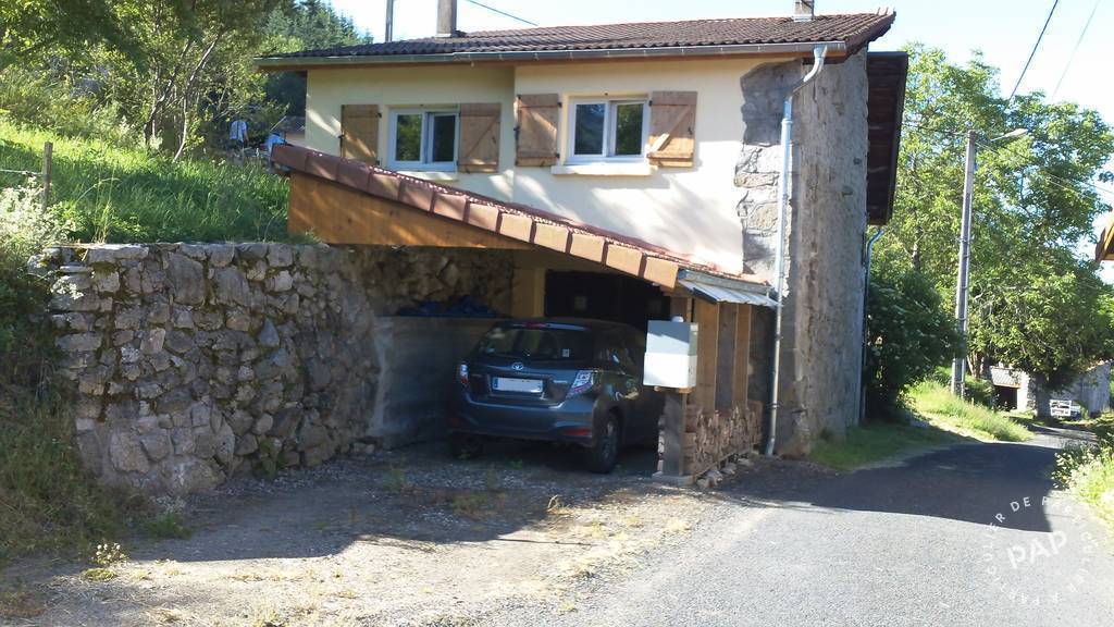 A rustic house with a stone facade is set against a green hillside. A covered parking area accommodates a car beneath the main entrance. Surrounding greenery enhances the rural charm, and a narrow road leads to the property.