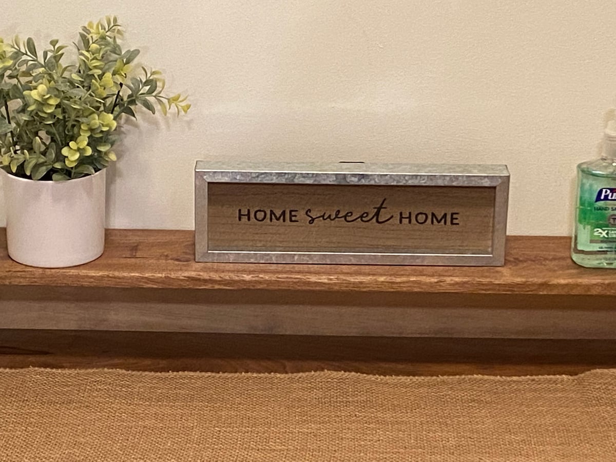 A wooden shelf displays a decorative sign reading 'HOME sweet HOME' with a galvanized metal frame. A small white pot holds greenery, complemented by a bottle of hand sanitizer nearby. The background features a neutral wall.