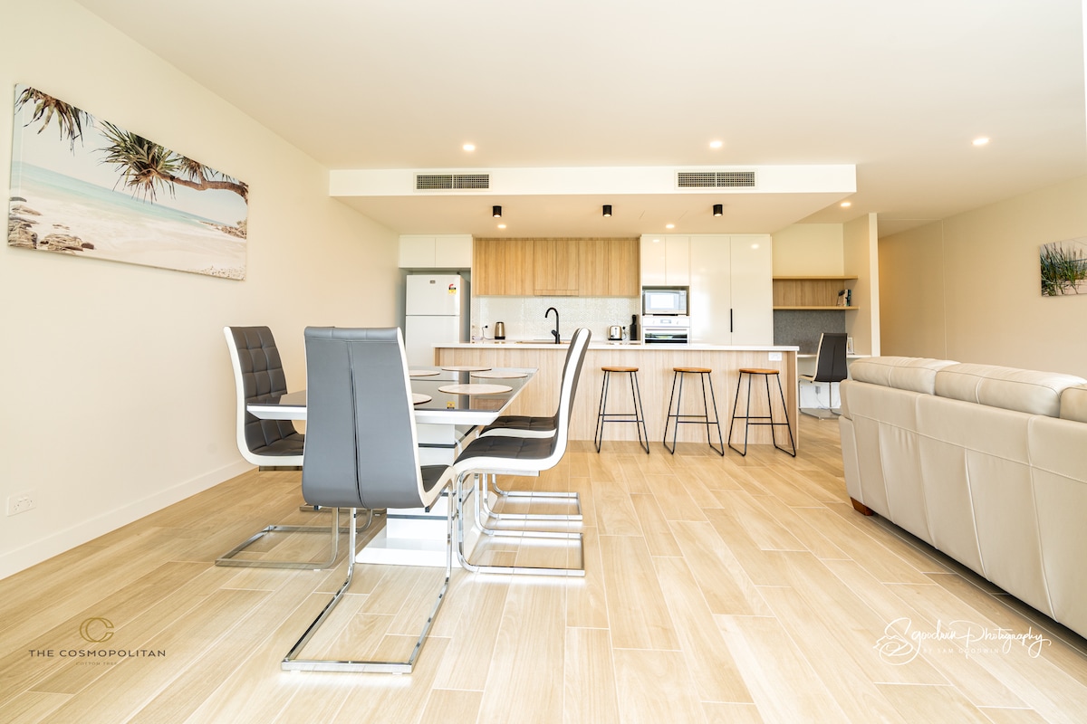 A spacious dining area features a modern glass table set for six, accompanied by sleek dining chairs. The open kitchen is equipped with contemporary appliances and light wood cabinetry, enhancing a clean and airy feel throughout the living space.