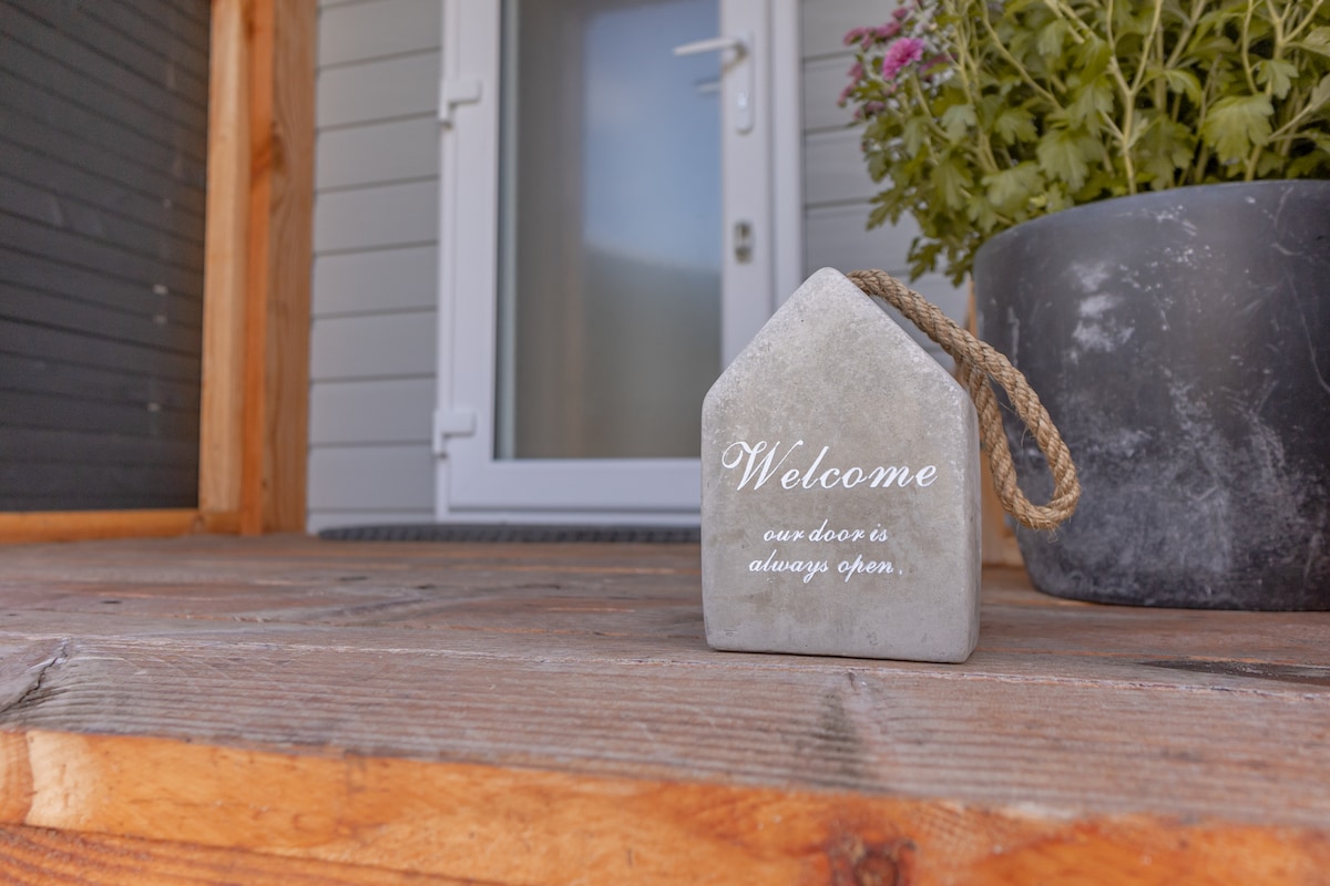 A stone welcome sign is positioned on a wooden porch, accompanied by a potted plant. The entrance door is visible in the background, enhancing the inviting ambiance of the space.
