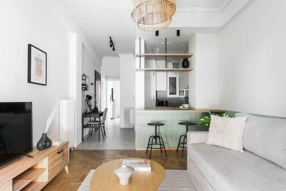 A bright and airy living area features a comfortable sofa and a wooden coffee table. The open-plan space connects to a modern kitchen with sleek cabinetry and bar seating. Natural light fills the room, highlighting the tasteful décor and warm wood accents.