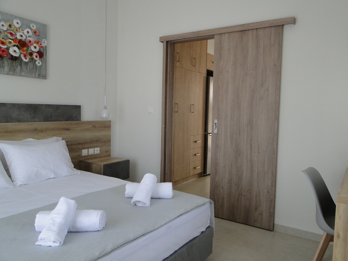 A spacious sleeping area features a comfortable bed with neatly arranged towels. The room includes a sliding wooden door leading to a closet, accompanied by a desk and chair. Natural light enters through a nearby window, enhancing the serene ambiance.