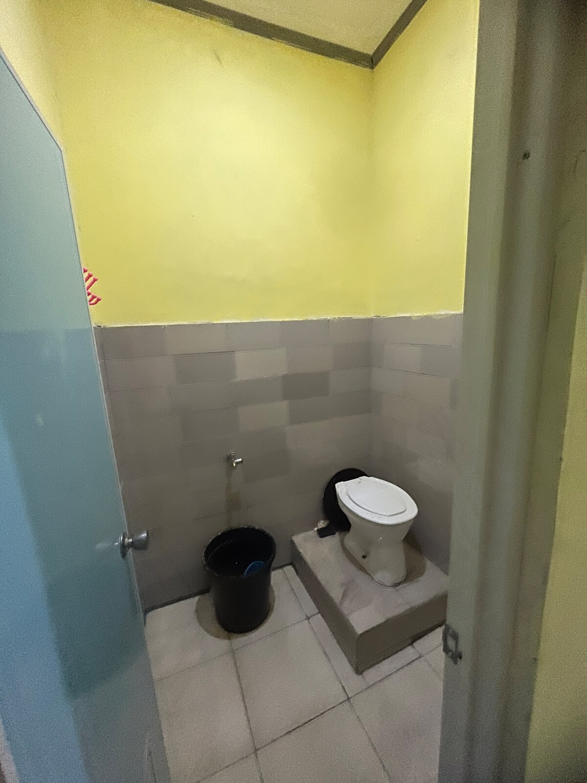 A compact bathroom is shown with a toilet positioned on a raised platform. The walls feature light yellow and gray tiles, providing a clean appearance. A trash bin is visible next to the toilet, and blue and gray ceramic tiles cover the floor.