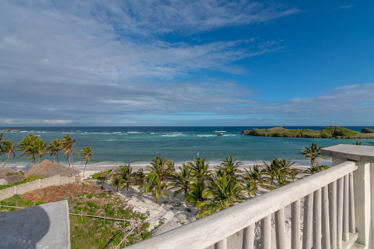 A panoramic view of the coastline is captured from a balcony, with gently swaying palm trees lining the sandy beach. The ocean sparkles under a clear blue sky, while distant islands are visible on the horizon.