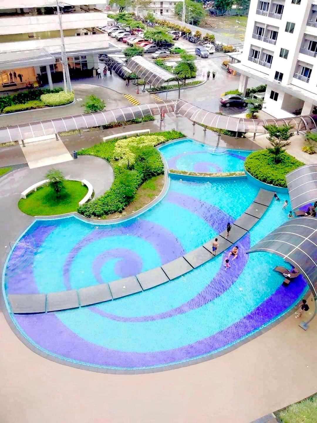 An aerial view of a swimming pool with a unique spiral design, featuring vibrant shades of blue. Surrounding greenery and walkways are visible, along with guests enjoying the water. Nearby, covered pathways connect to various areas of the property.