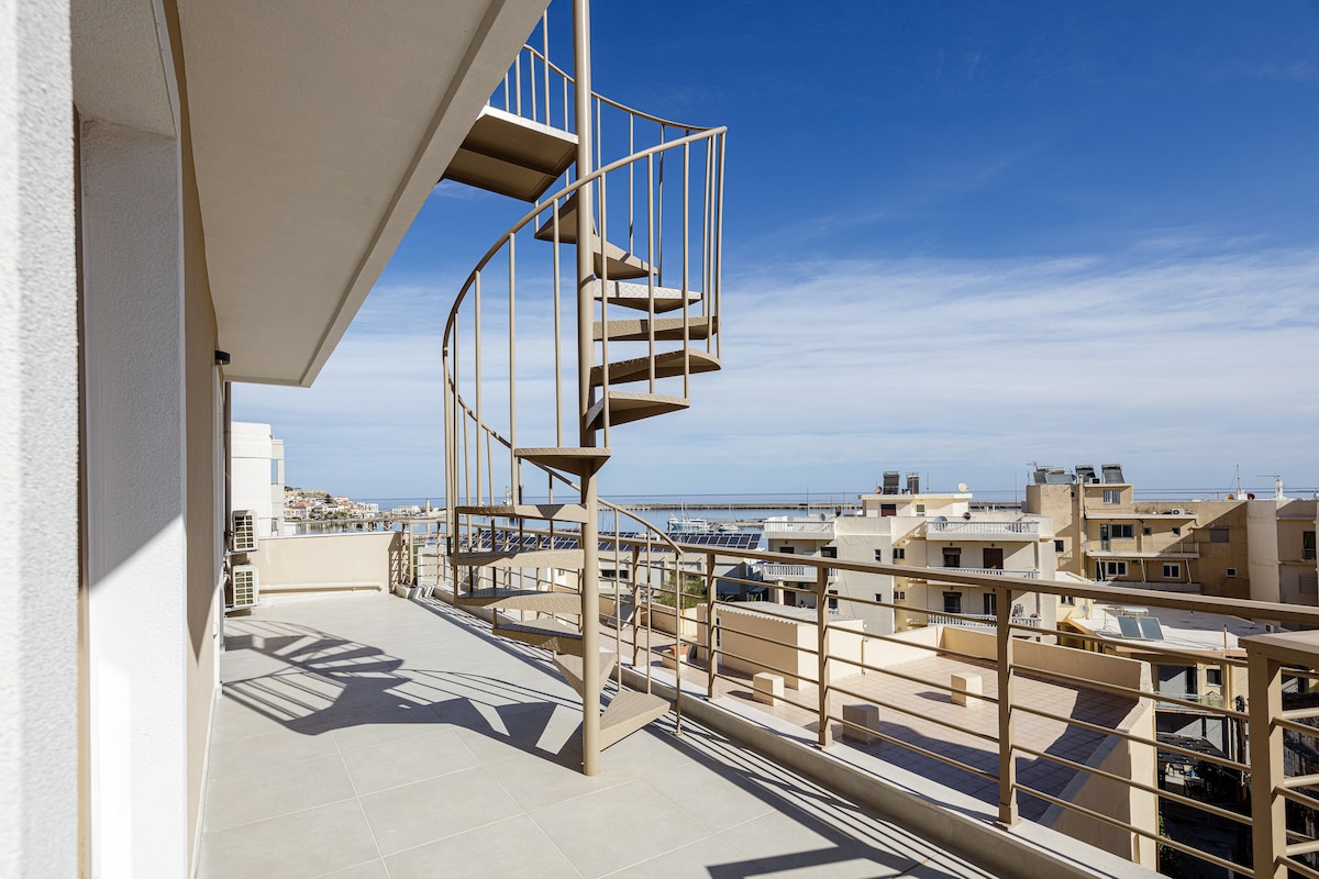 A spiral staircase leads from the spacious rooftop terrace, enhancing access to panoramic views of the surrounding buildings and the sea. The terrace is designed with safety railings and features a light-toned tiled floor that reflects natural sunlight.
