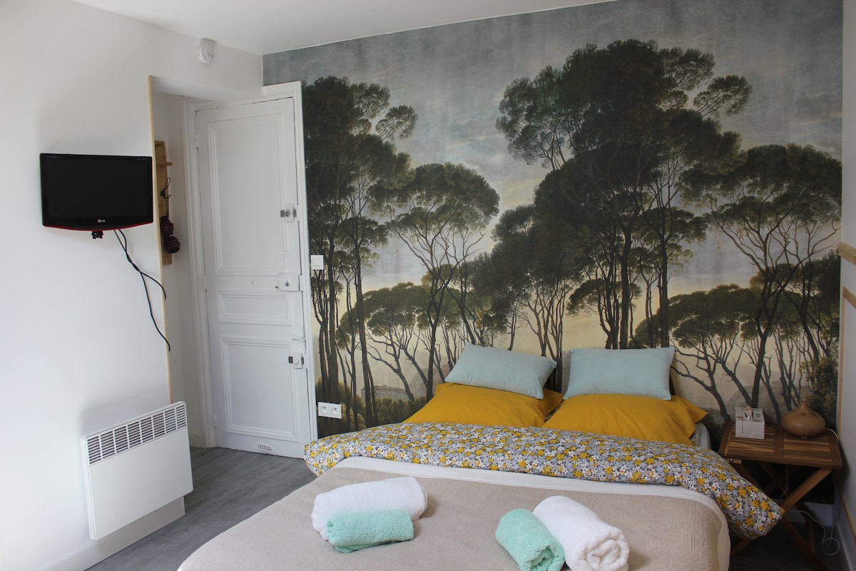 A cozy sleeping area features a spacious bed adorned with yellow and blue pillows. The wall behind showcases a serene mural of trees. A large blanket sits at the foot of the bed, with neatly folded towels placed on top.