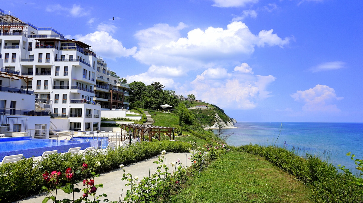 The image showcases the modern exterior of the Biala Cliff complex, featuring white buildings nestled along the coastline. A large infinity pool reflects the blue sky, while manicured greenery and rose bushes enhance the scenic landscape. The tranquil sea can be seen in the background.