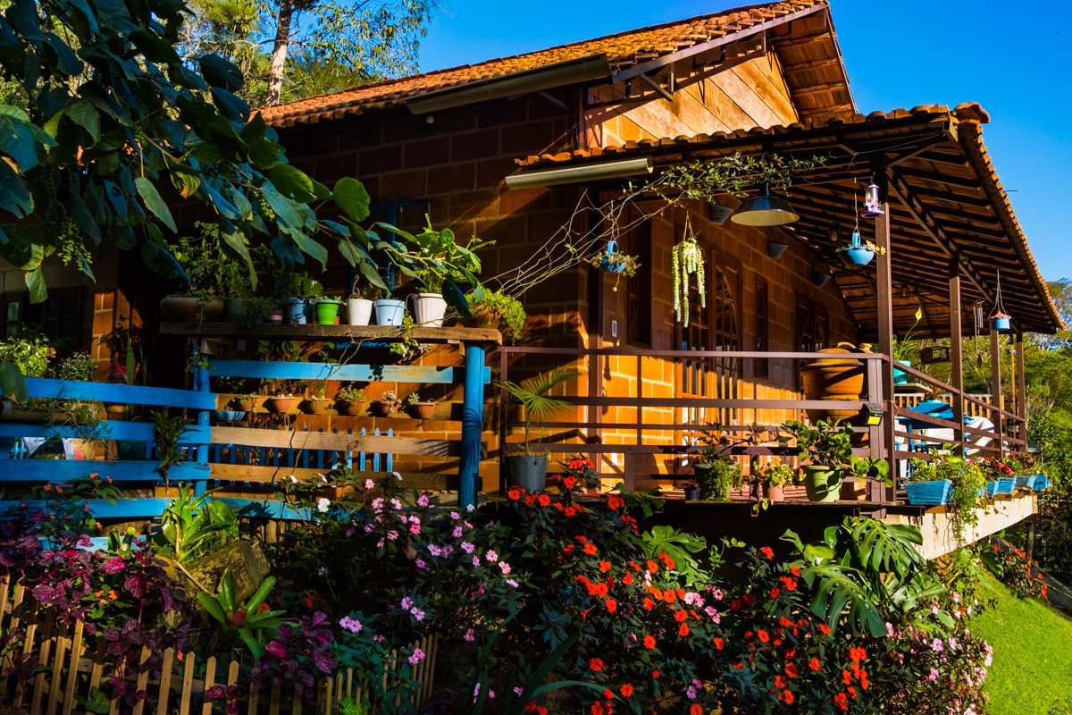 A charming house is set against a clear blue sky, with wooden architecture featuring a porch adorned with colorful plants and hanging decorations. Lush flowers in various hues create a vibrant border around the property, emphasizing the connection with nature.