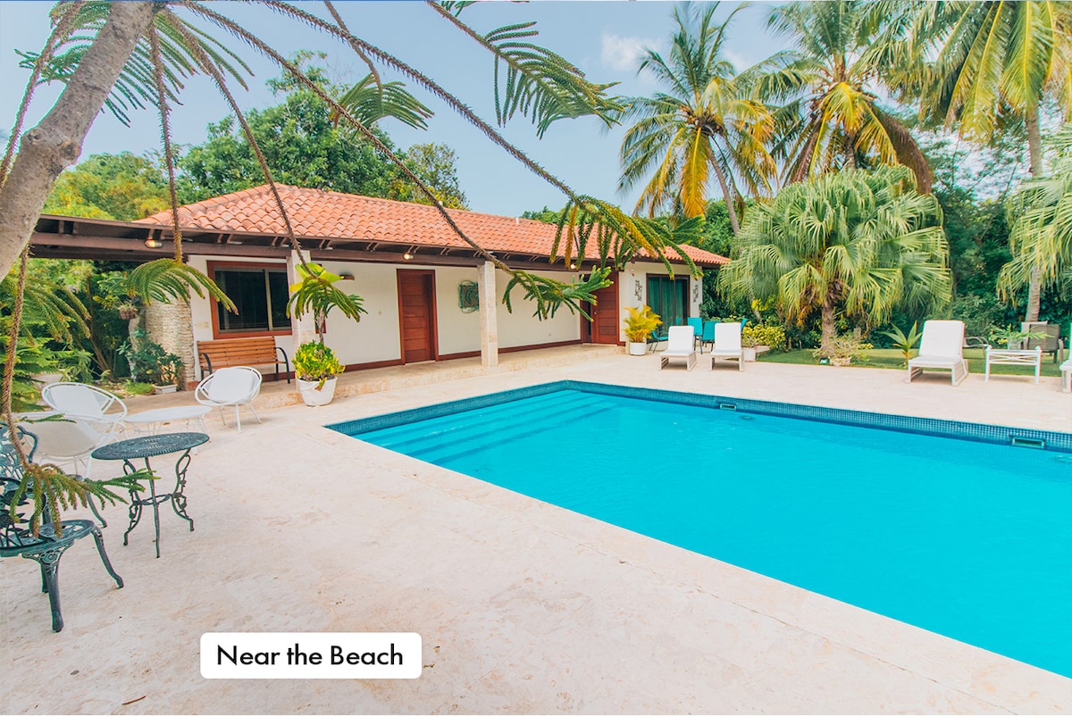 An outdoor area features a clear blue swimming pool surrounded by a stone deck. Lush palm trees and tropical plants border the space, while several white lounge chairs are positioned nearby. The villa's exterior is visible, showcasing a tiled roof and wooden doors.