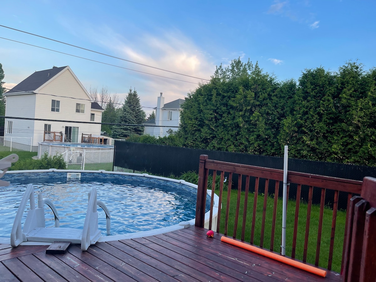 A spacious wooden deck surrounds a circular pool, set in a grassy backyard. Lush greenery lines the perimeter, providing privacy. Nearby houses enhance the neighborhood feel, framed by a clear blue sky streaked with soft clouds.